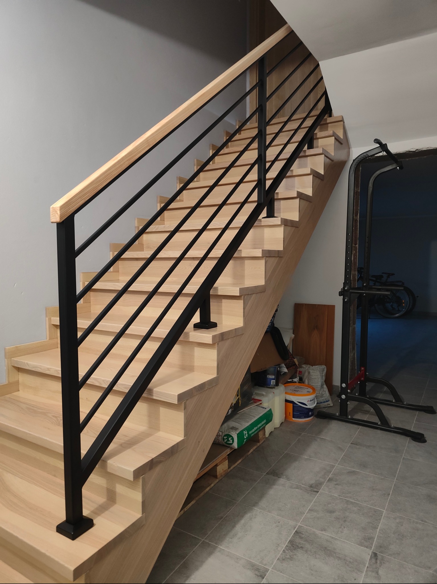 Jasne drewniane schody z czarną metalową balustradą i drewnianą poręczą, widok z dołu. W tle widoczne akcesoria do ćwiczeń i materiały budowlane.