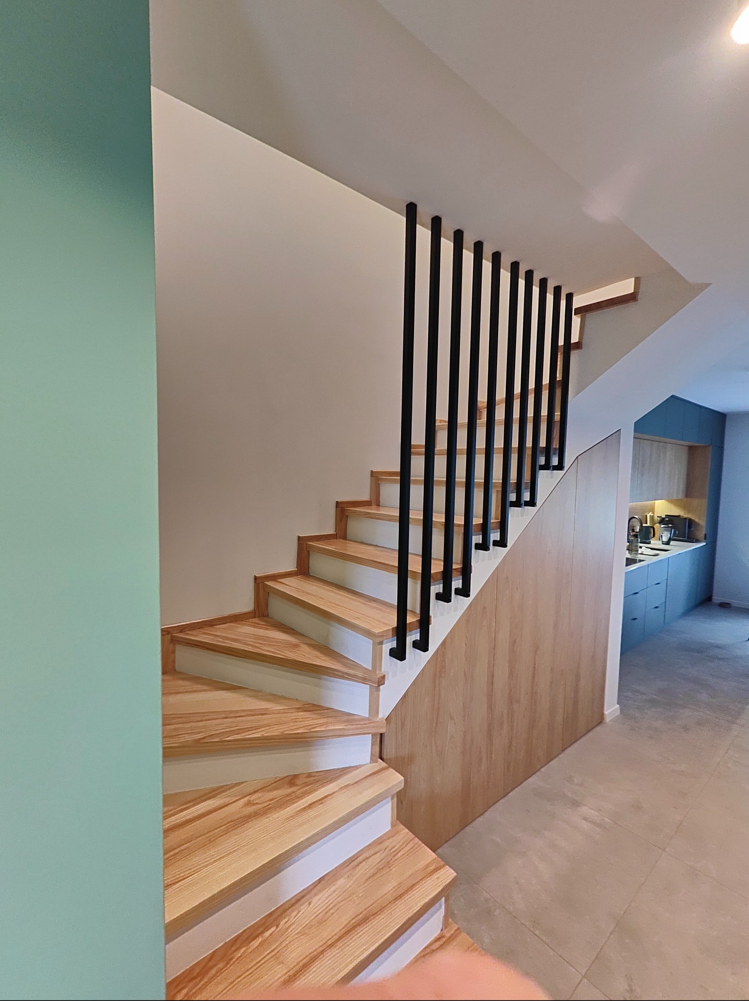 Nowoczesne schody drewniane z czarną, minimalistyczną balustradą. Jasne drewno stopni kontrastuje z bielą podstopnic i ścianą w tle. Widok na kuchnię w głębi.