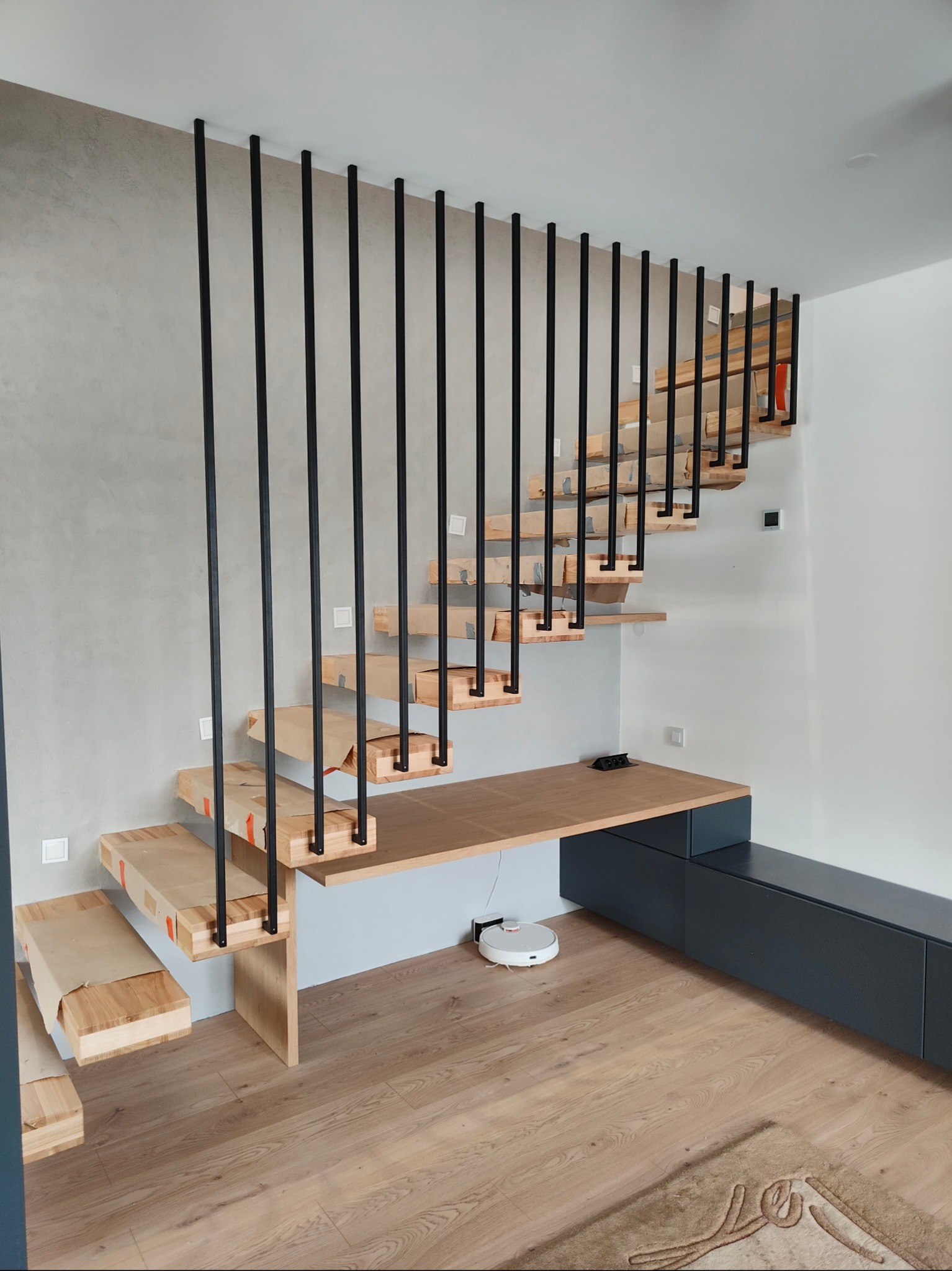 Nowoczesne schody drewniane z czarnymi, metalowymi balustradami pionowymi. Stopnie zabezpieczone folią, pod schodami biurko i szafka w kolorze grafitowym.