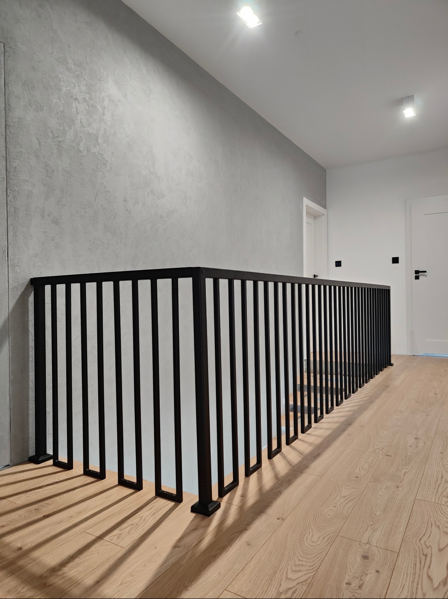 Nowoczesna, czarna balustrada metalowa z pionowymi szczeblami na jasnym parkiecie. Szara ściana z efektem dekoracyjnym w tle. Minimalistyczny design, oświetlenie sufitowe.