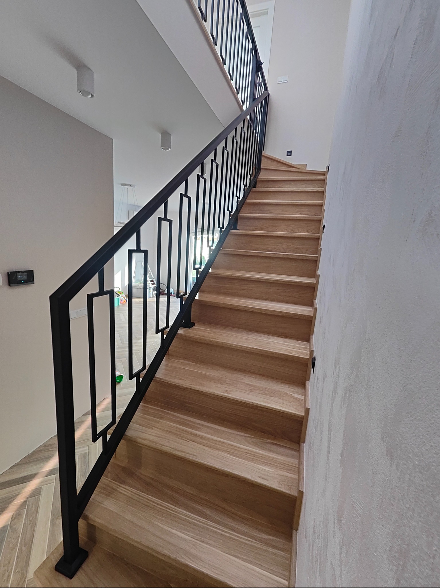 Drewniane schody z czarną, metalową balustradą o geometrycznym wzorze. Widok z dołu, podkreślający solidne wykonanie i nowoczesny design.