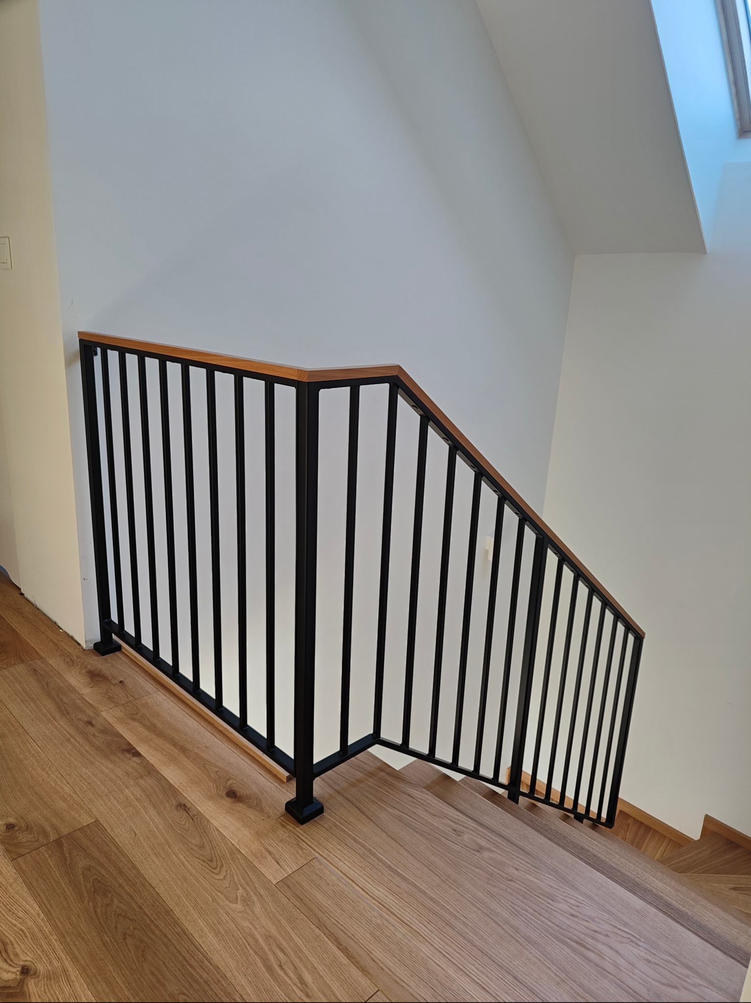 Nowoczesne poręcze schodowe z czarnych, metalowych prętów i drewnianego pochwytu, zamontowane na drewnianych schodach w jasnym wnętrzu.
