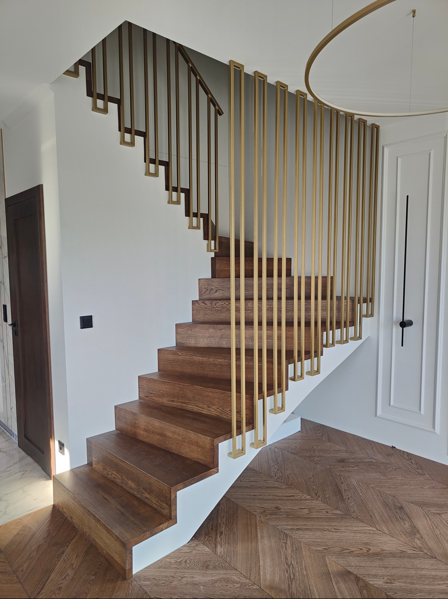 Drewniane schody z złotymi, pionowymi elementami balustrady w Perzowie. Parkiet ułożony w jodełkę. Minimalistyczny design, eleganckie wykończenie.