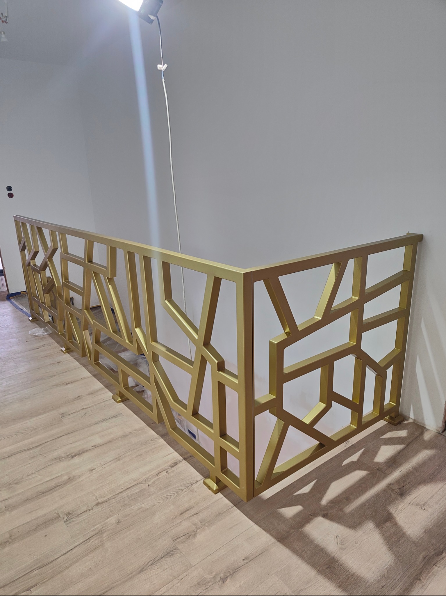 Złota balustrada wewnętrzna z geometrycznym wzorem, zamontowana na jasnej podłodze. Nowoczesny design, oświetlenie górne podkreśla metaliczną fakturę.