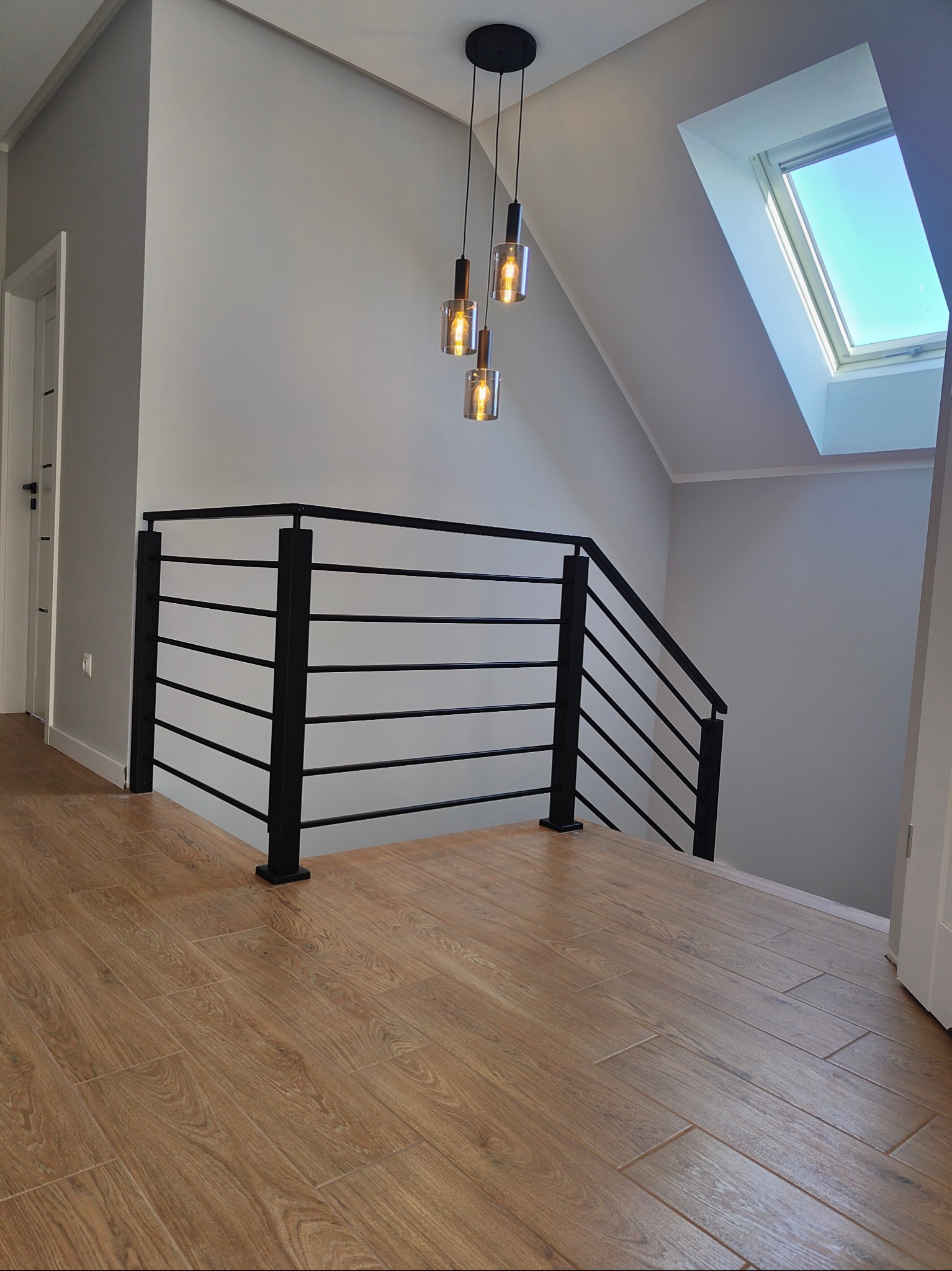 Nowoczesna, czarna balustrada schodowa z poziomymi prętami, oświetlona wiszącymi lampami. Ujęcie z perspektywy podłogi z drewnopodobnych paneli, widoczne okno dachowe.