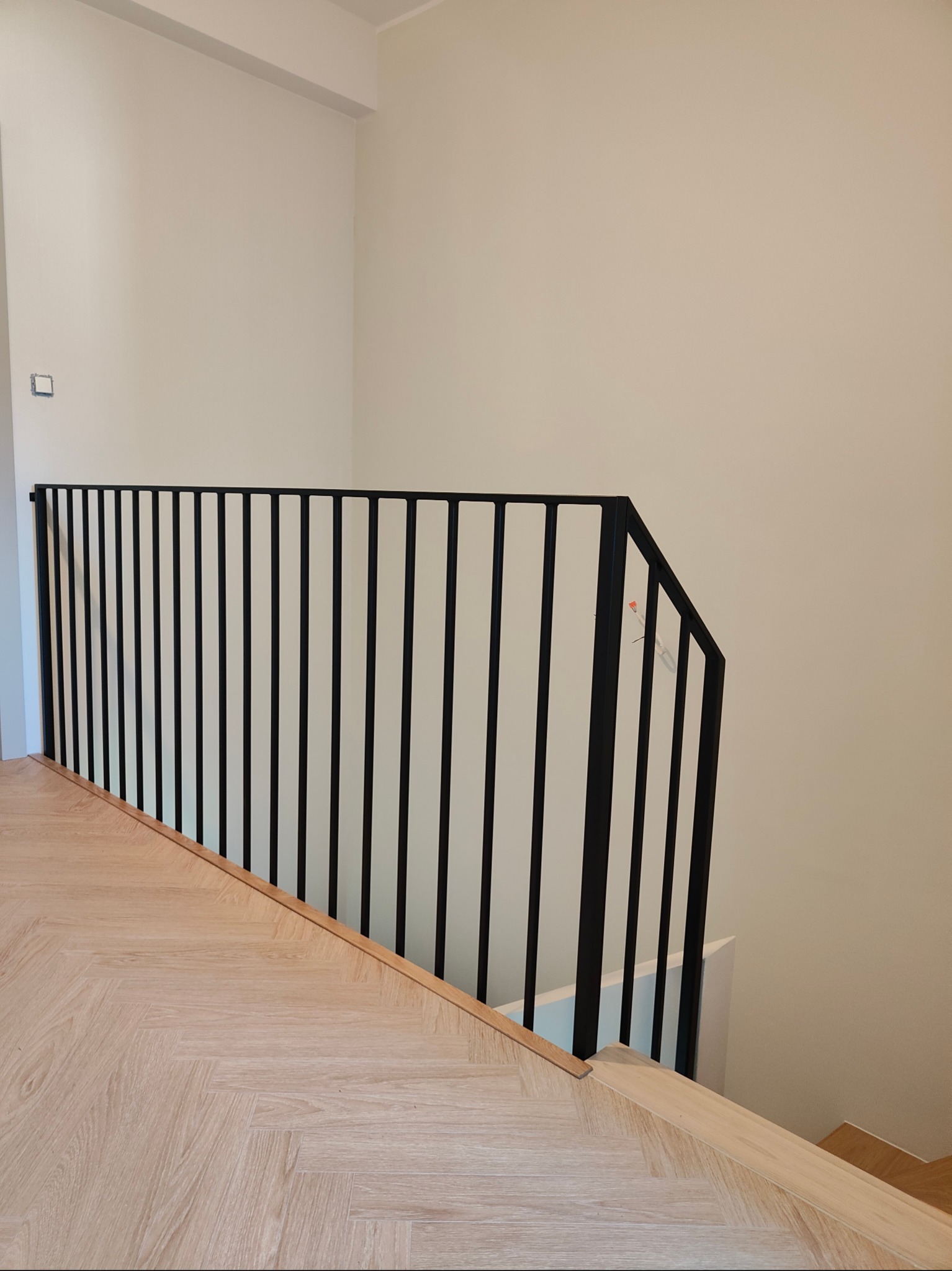 Minimalistyczna, czarna balustrada schodowa z pionowych prętów, zamontowana na drewnianych schodach w jasnym wnętrzu. Widoczne detale łączenia balustrady.