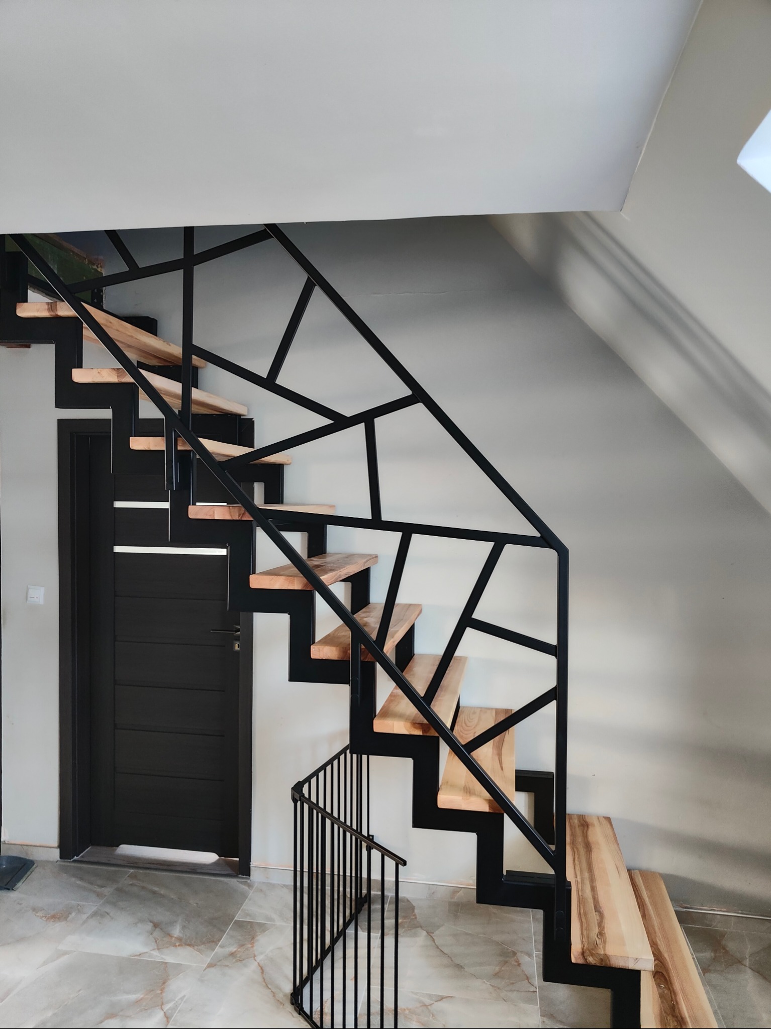 Nowoczesne schody z drewnianymi stopniami i czarną, geometryczną balustradą metalową. Widok z dołu, marmurowa podłoga i czarne drzwi w tle.