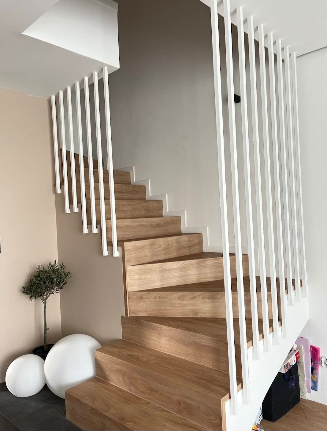 Drewniane schody z nowoczesną, białą balustradą z pionowych prętów. U dołu schodów dekoracyjne, białe kule i małe drzewko w doniczce. Minimalistyczny design.