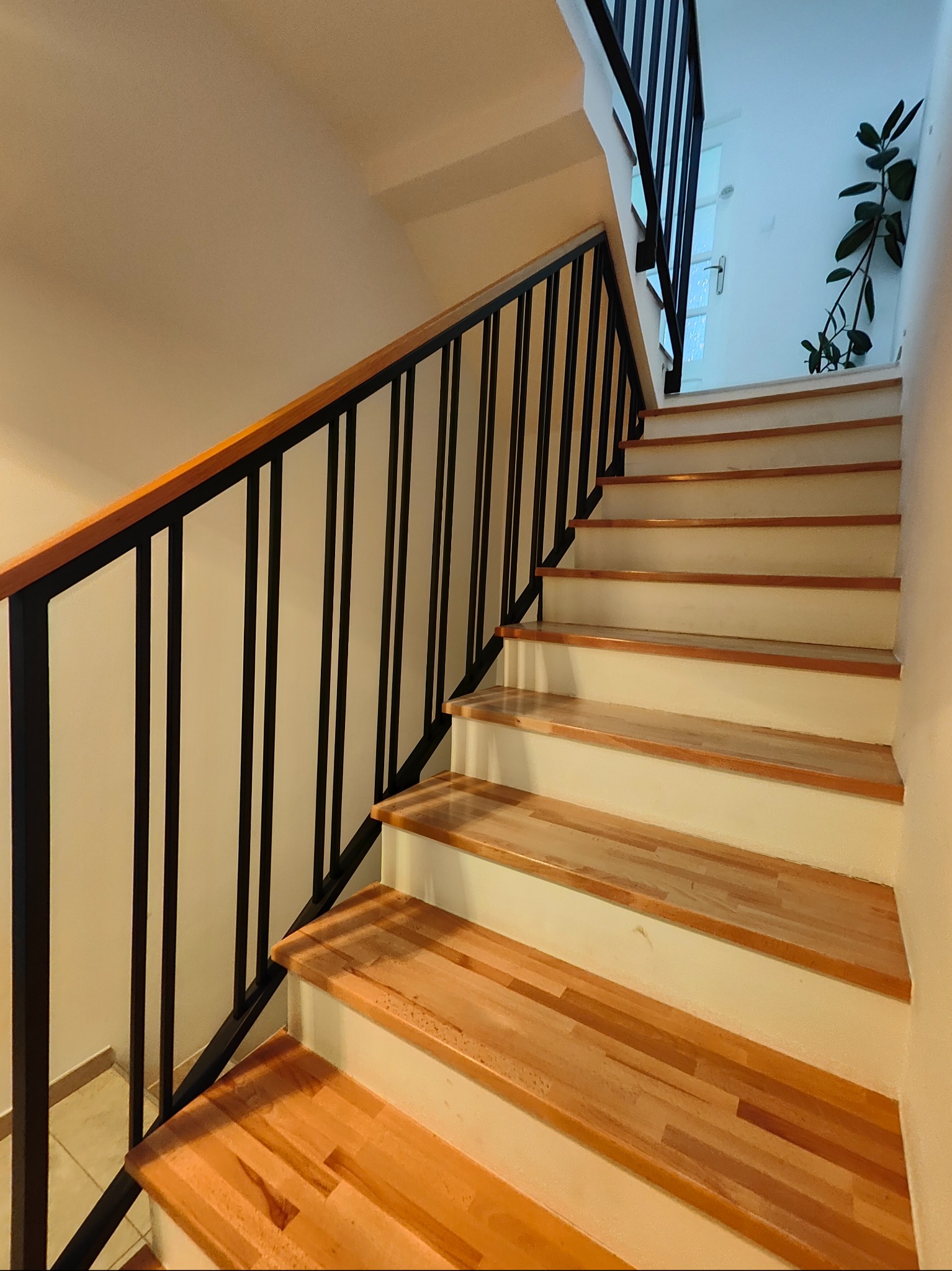 Schody z drewnianymi stopniami i czarną, metalową balustradą z drewnianym poręczem. Widok z dołu schodów, w tle roślina doniczkowa i okno.