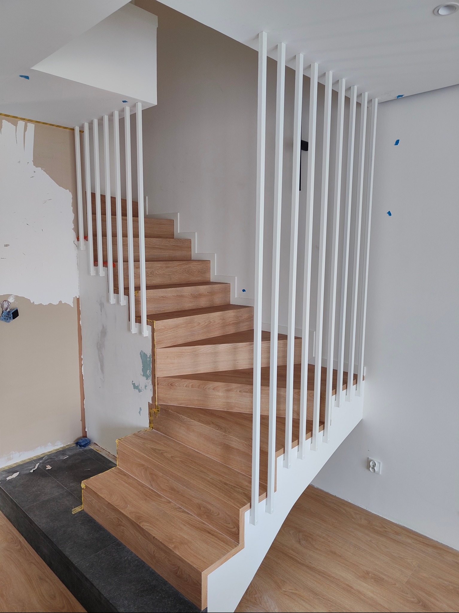 Drewniane schody spiralne z białymi, pionowymi balustradami w nowoczesnym wnętrzu. Widoczne niedokończone prace wykończeniowe na ścianie i podłodze.