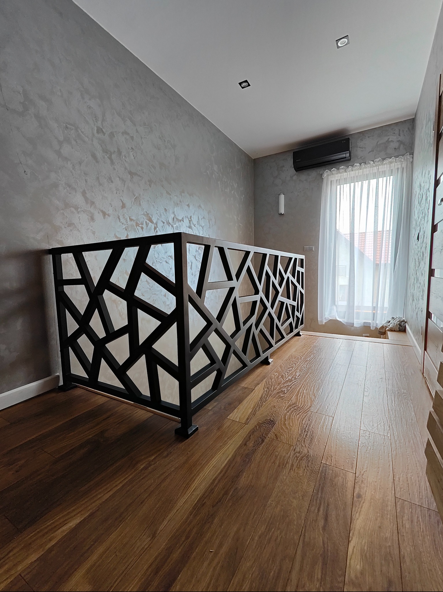 Nowoczesna balustrada metalowa z geometrycznym wzorem w czarnym kolorze, zamontowana w jasnym wnętrzu z drewnianą podłogą i dekoracyjną ścianą.
