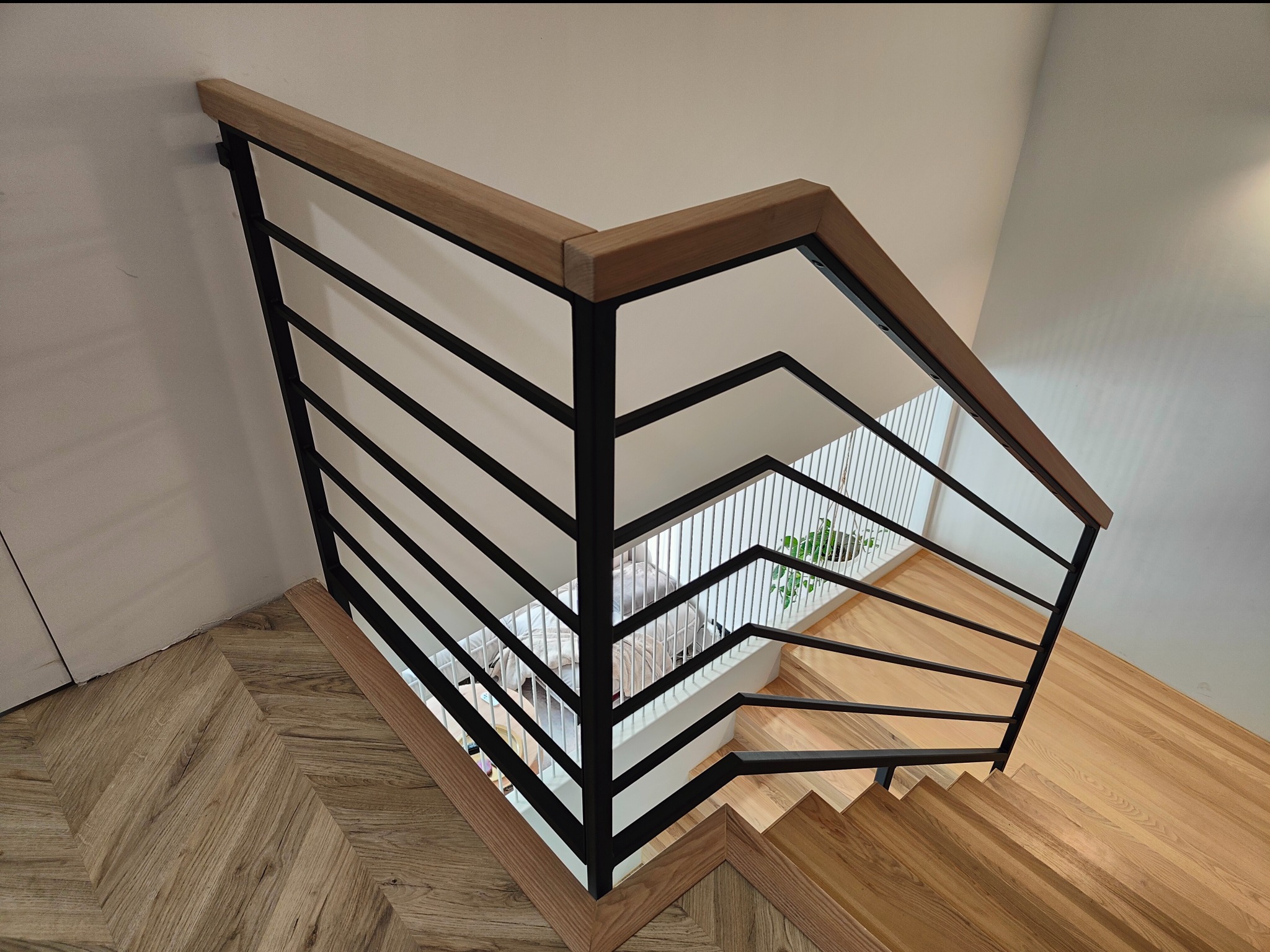 Nowoczesne schody z czarną metalową balustradą i drewnianym poręczem, widok z góry. Parkiet w jodełkę na podeście, jasne drewniane stopnie. Minimalistyczny design.