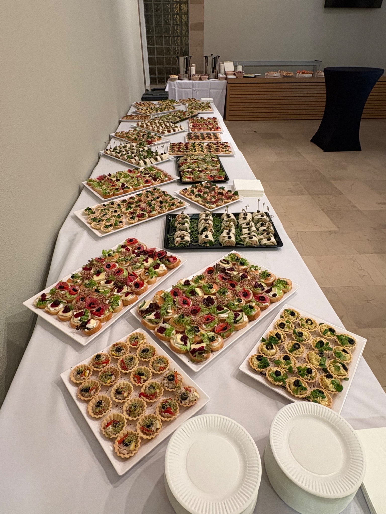 Długi stół cateringowy z różnorodnymi przekąskami: tartaletki, kanapki z warzywami i mięsem, roladki. Biały obrus, talerzyki. Neutralne tło.