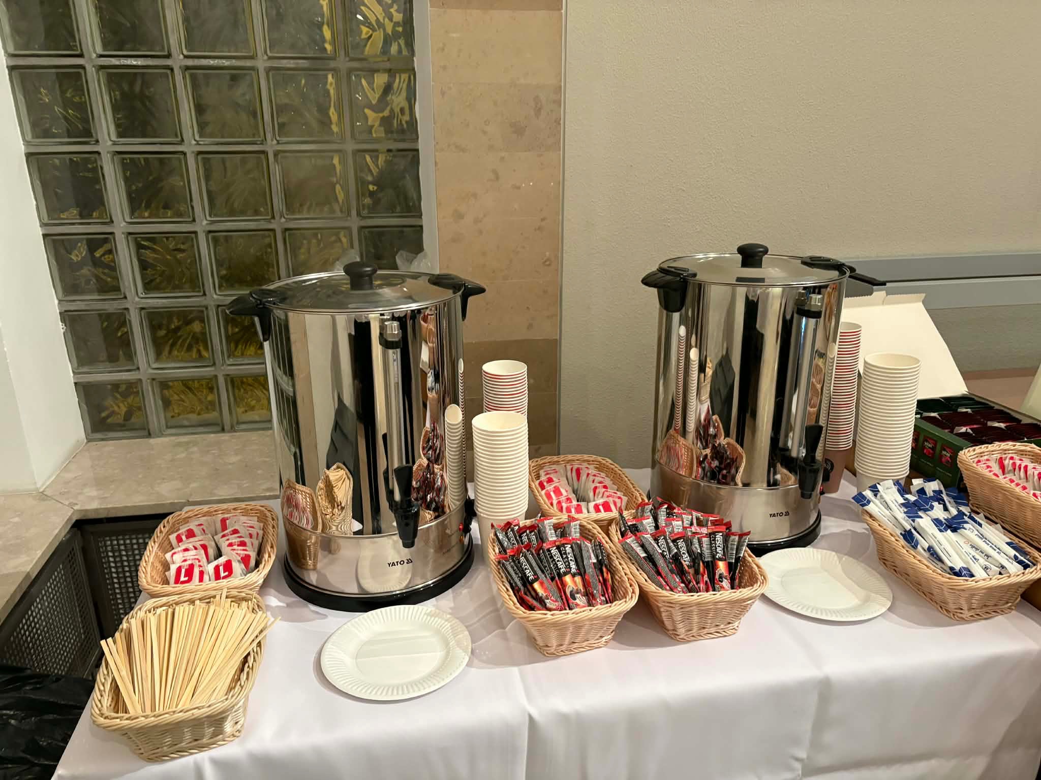 Stół cateringowy z dwoma dużymi termosami, koszykami z kawą, cukrem i mieszadełkami, oraz stosem papierowych kubków i talerzyków. Przygotowany serwis kawowy.