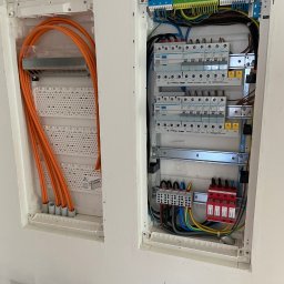 ELEKTRO-HERBUT Marek Herbut - Dwa panele elektryczne w ścianie: pomarańczowe kable w lewym panelu, bezpieczniki i okablowanie w prawym. Białe obudowy, szare tło ściany.