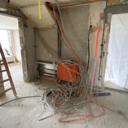 ELEKTRO-HERBUT Marek Herbut - Rozdzielnia elektryczna z przewodami w surowym wnętrzu. Widoczne kable, puszki i drabina. Ściany w trakcie wykończenia, surowy beton. Etap prac instalacyjnych.