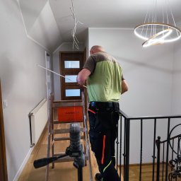 ELEKTRO-HERBUT Marek Herbut - Elektryk na drabinie, poziomuje laserowo sufit pod montaż nowoczesnej lampy wiszącej w dwupoziomowym domu. Widoczne kable i narzędzia.