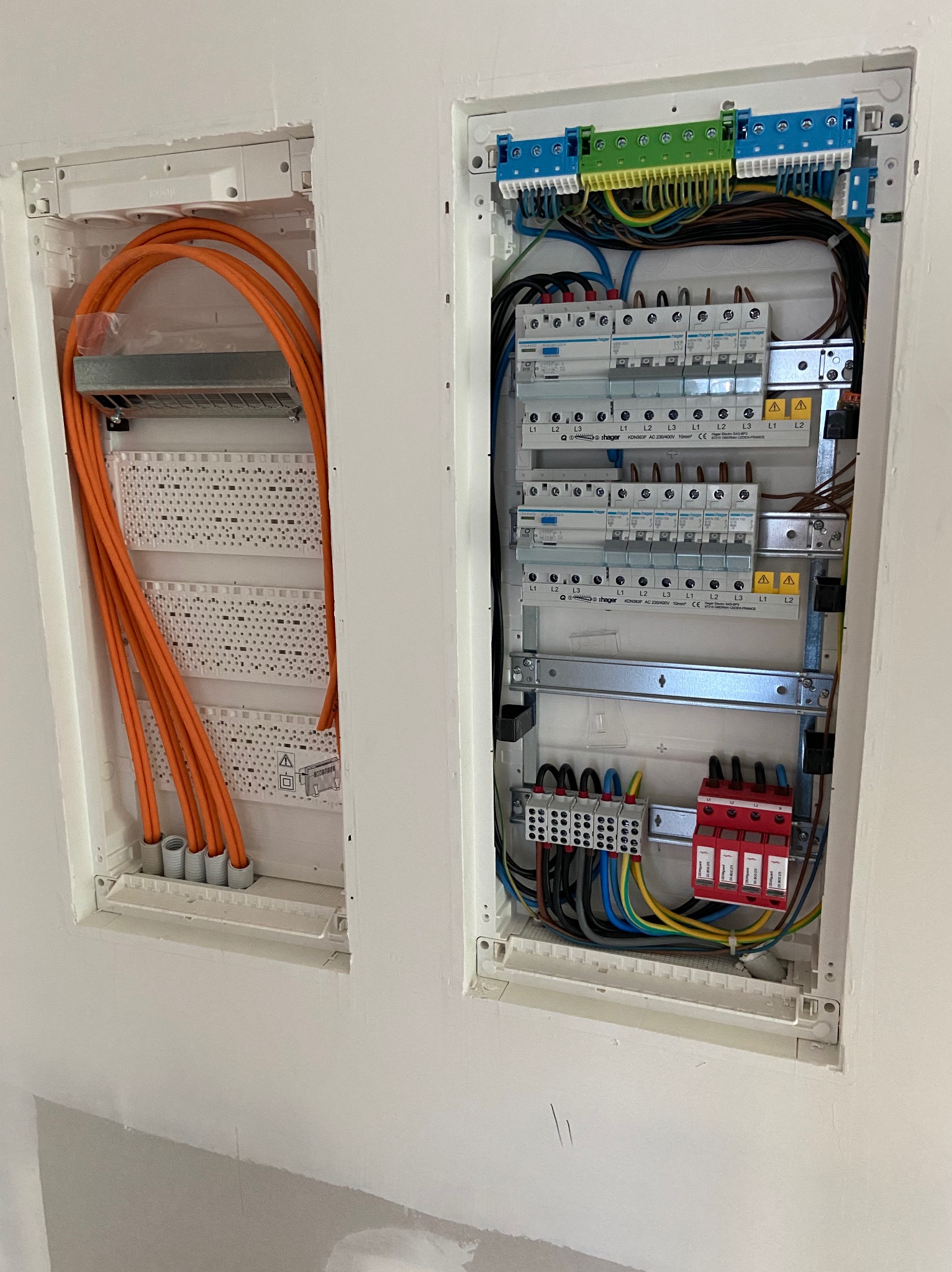 Dwa panele elektryczne w ścianie: pomarańczowe kable w lewym panelu, bezpieczniki i okablowanie w prawym. Białe obudowy, szare tło ściany.