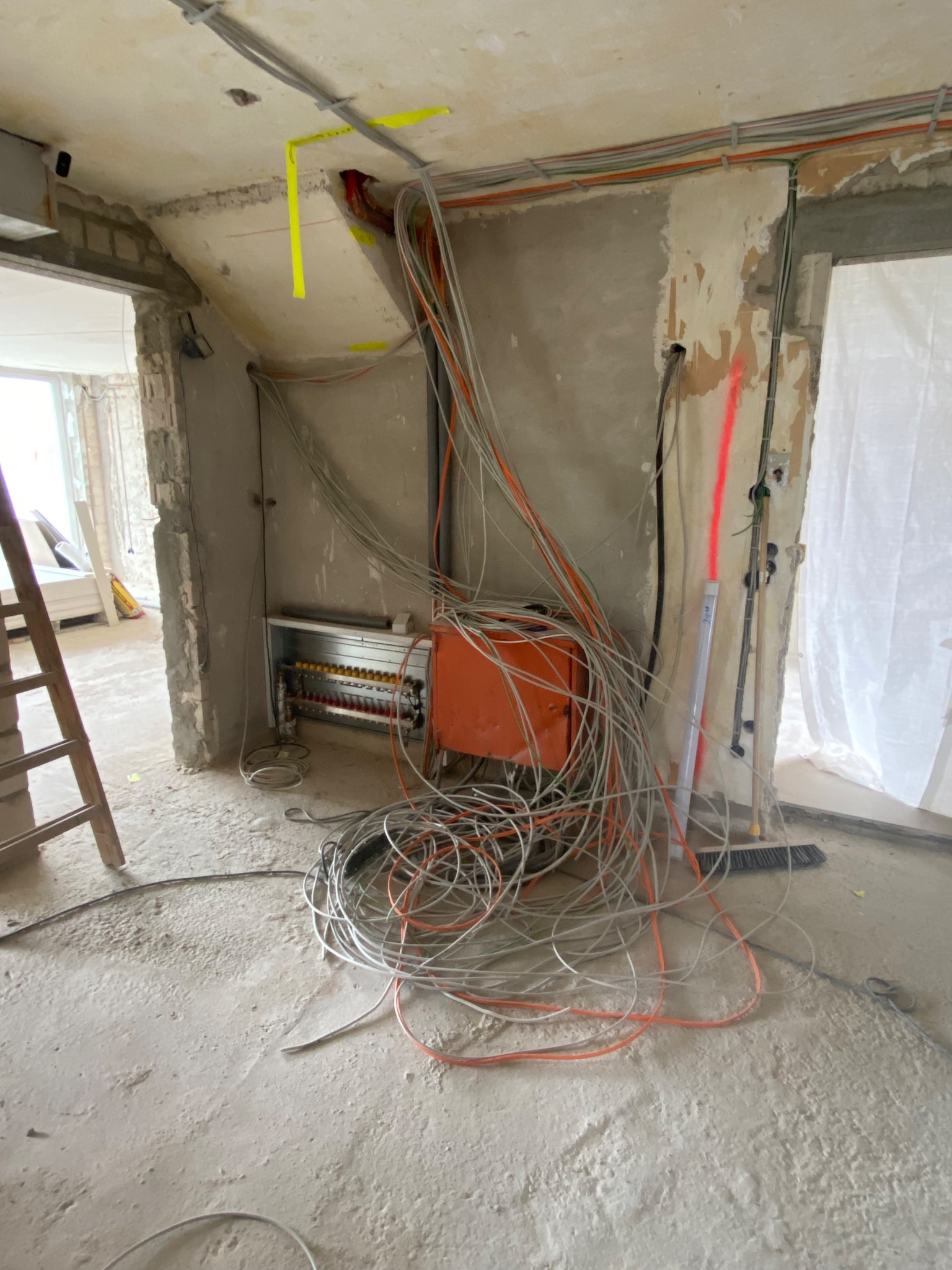 Rozdzielnia elektryczna z przewodami w surowym wnętrzu. Widoczne kable, puszki i drabina. Ściany w trakcie wykończenia, surowy beton. Etap prac instalacyjnych.