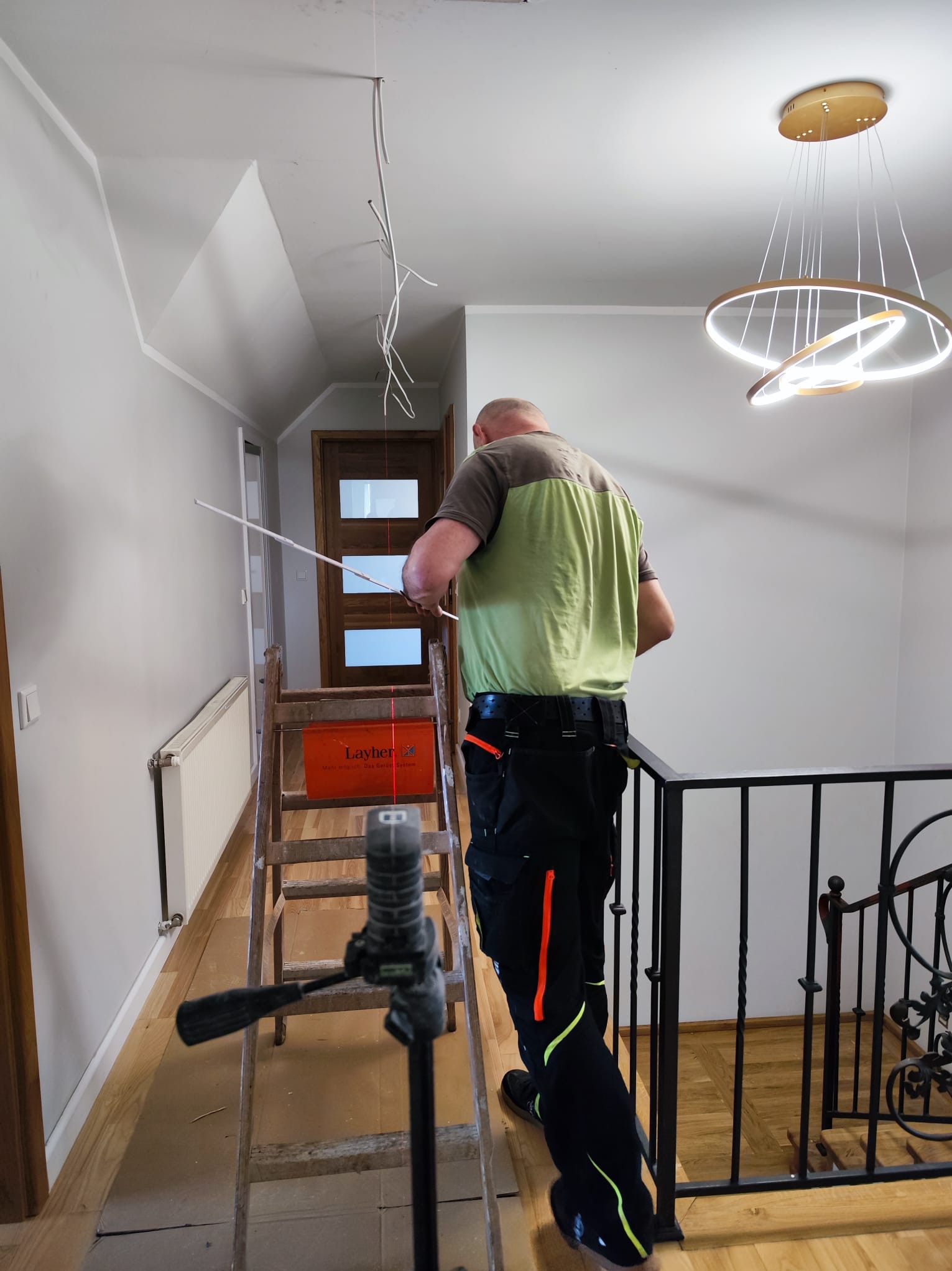 Elektryk na drabinie, poziomuje laserowo sufit pod montaż nowoczesnej lampy wiszącej w dwupoziomowym domu. Widoczne kable i narzędzia.