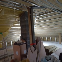 Reno-j - Poddasze w trakcie adaptacji: widoczne ocieplenie wełną mineralną, konstrukcja sufitu podwieszanego z profili metalowych, komin z cegły i materiały budowlane.