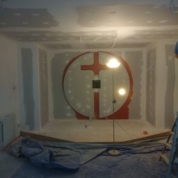 Reno-j - Pokój w trakcie prac wykończeniowych: płyty karton-gips na ścianach, okrągła dekoracja z krzyżem w tle, podłoga zabezpieczona folią malarską, widoczna lampa stojąca.