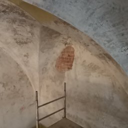 Reno-j - Klatka schodowa w trakcie remontu, widoczna ściana z fragmentem odsłoniętej cegły i metalowe rusztowanie. W tle sufit z widocznymi śladami wilgoci.
