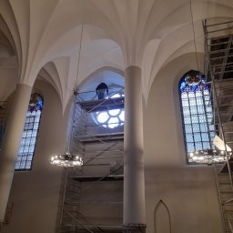 Reno-j - Wnętrze kościoła w trakcie renowacji: rusztowania, witraże, kolumny i sklepienie łukowe. Widoczny pracownik na rusztowaniu przy okrągłym oknie.