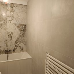 Wykończona łazienka z wanną, grzejnikiem drabinkowym i płytkami imitującymi marmur na ścianie. Widoczna żarówka na kablu. Minimalistyczny design.