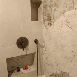 Wykończona łazienka z beżowymi i marmurowymi płytkami, wnękami w ścianie, baterią prysznicową w kolorze ciemnego brązu i białą wanną. Widok z bliska na detale.