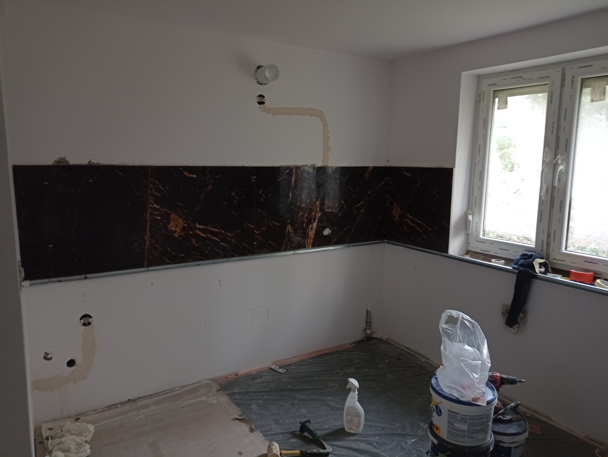 Częściowo wykończona kuchnia z czarnymi panelami ściennymi imitującymi marmur, widoczne narzędzia i materiały budowlane, okno w tle. Etap prac wykończeniowych.