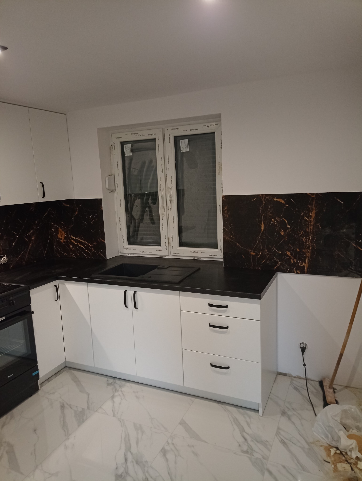 Nowoczesna kuchnia z białymi szafkami, czarnym blatem i marmurową okładziną ściany. Widoczne okno i fragment podłogi z marmurowym wzorem. Minimalistyczny design.