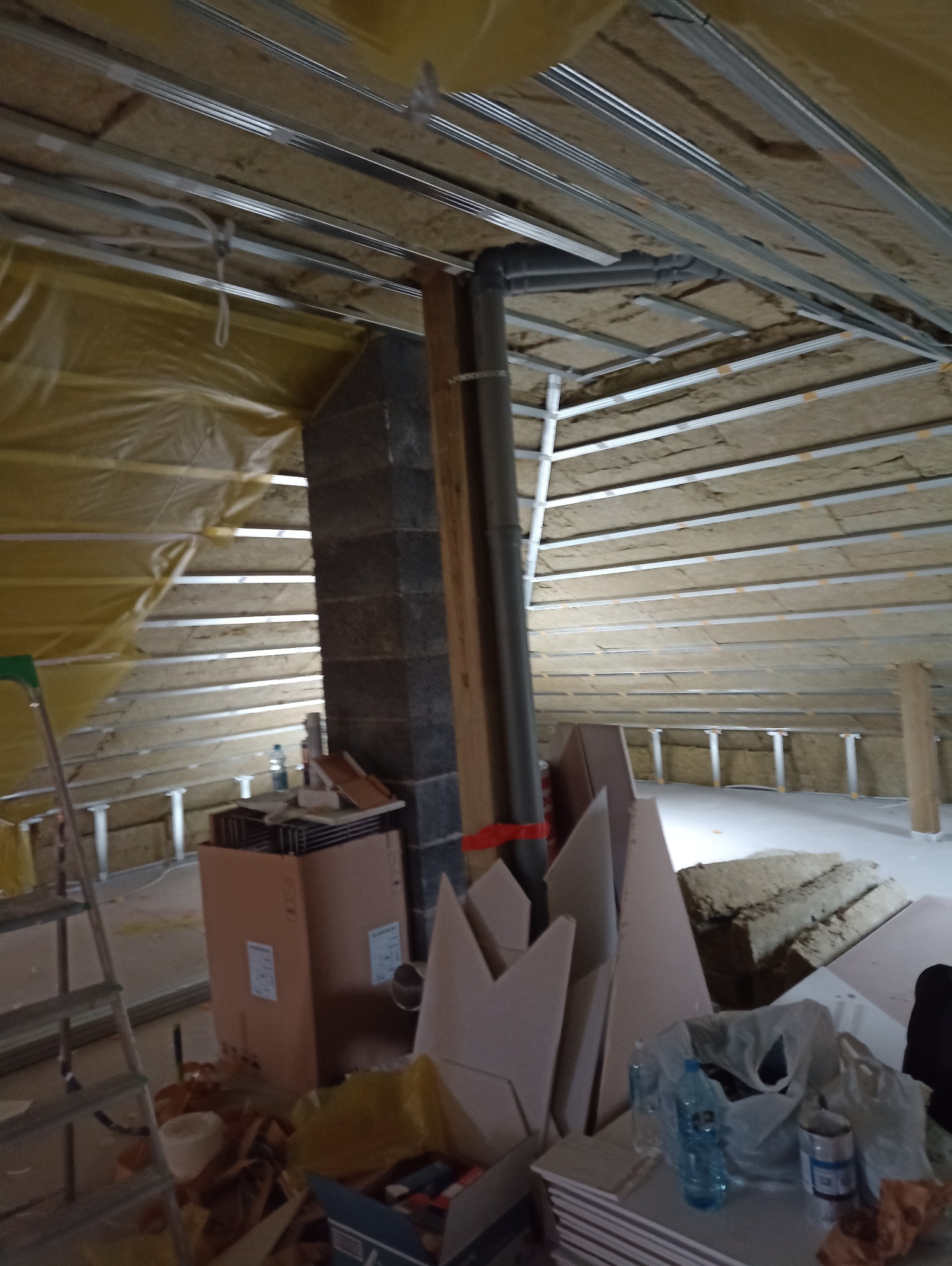 Poddasze w trakcie adaptacji: widoczne ocieplenie wełną mineralną, konstrukcja sufitu podwieszanego z profili metalowych, komin z cegły i materiały budowlane.