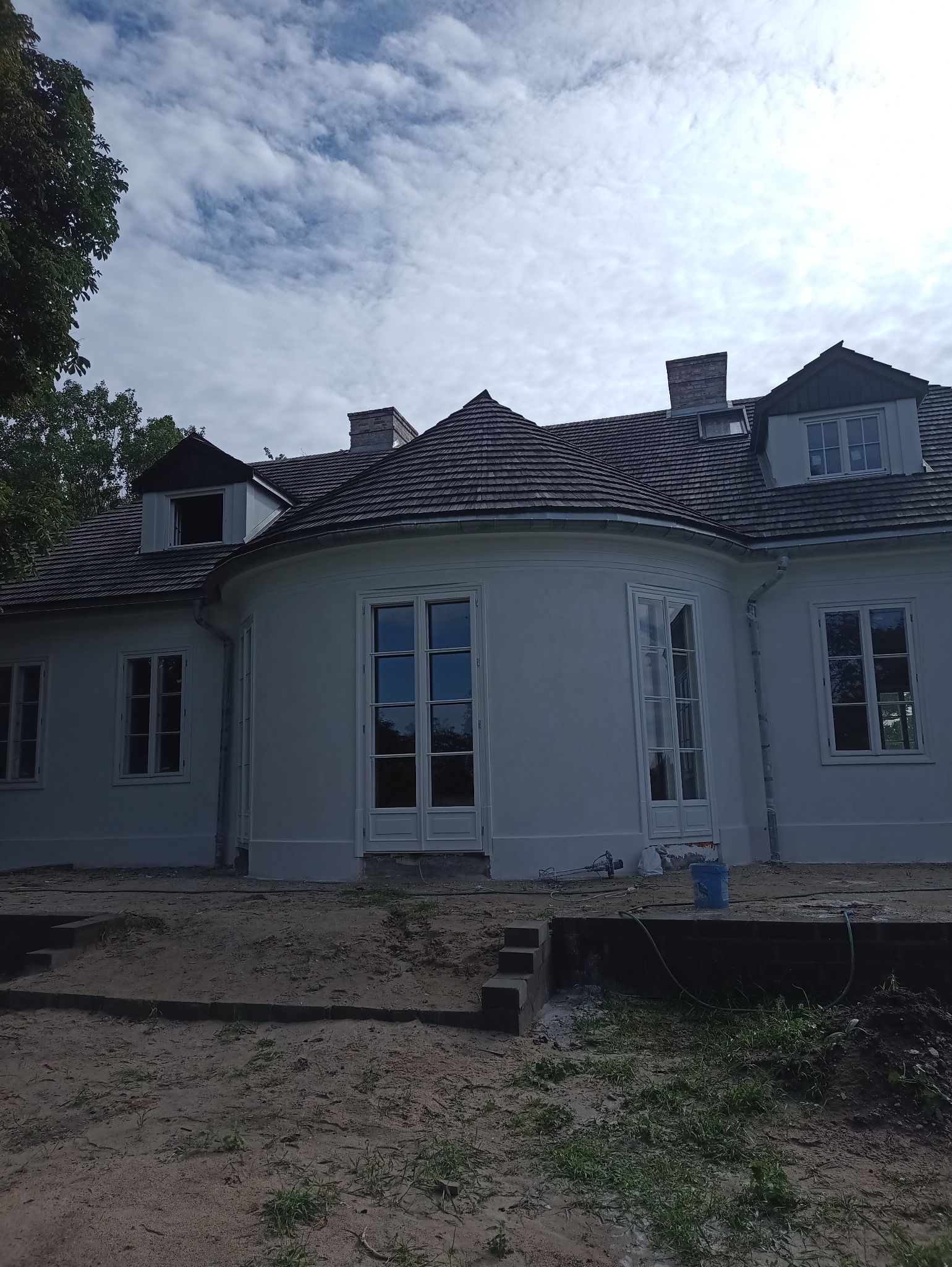 Biały, odnowiony budynek z szarą dachówką i okrągłą częścią. Widoczne ślady prac remontowych na ziemi. Błękitne niebo z chmurami w tle.