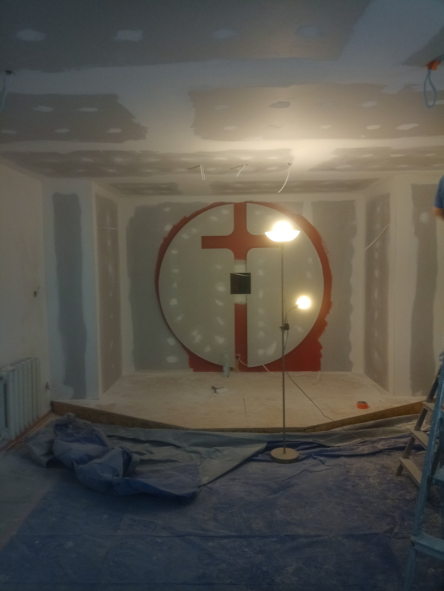 Pokój w trakcie prac wykończeniowych: płyty karton-gips na ścianach, okrągła dekoracja z krzyżem w tle, podłoga zabezpieczona folią malarską, widoczna lampa stojąca.