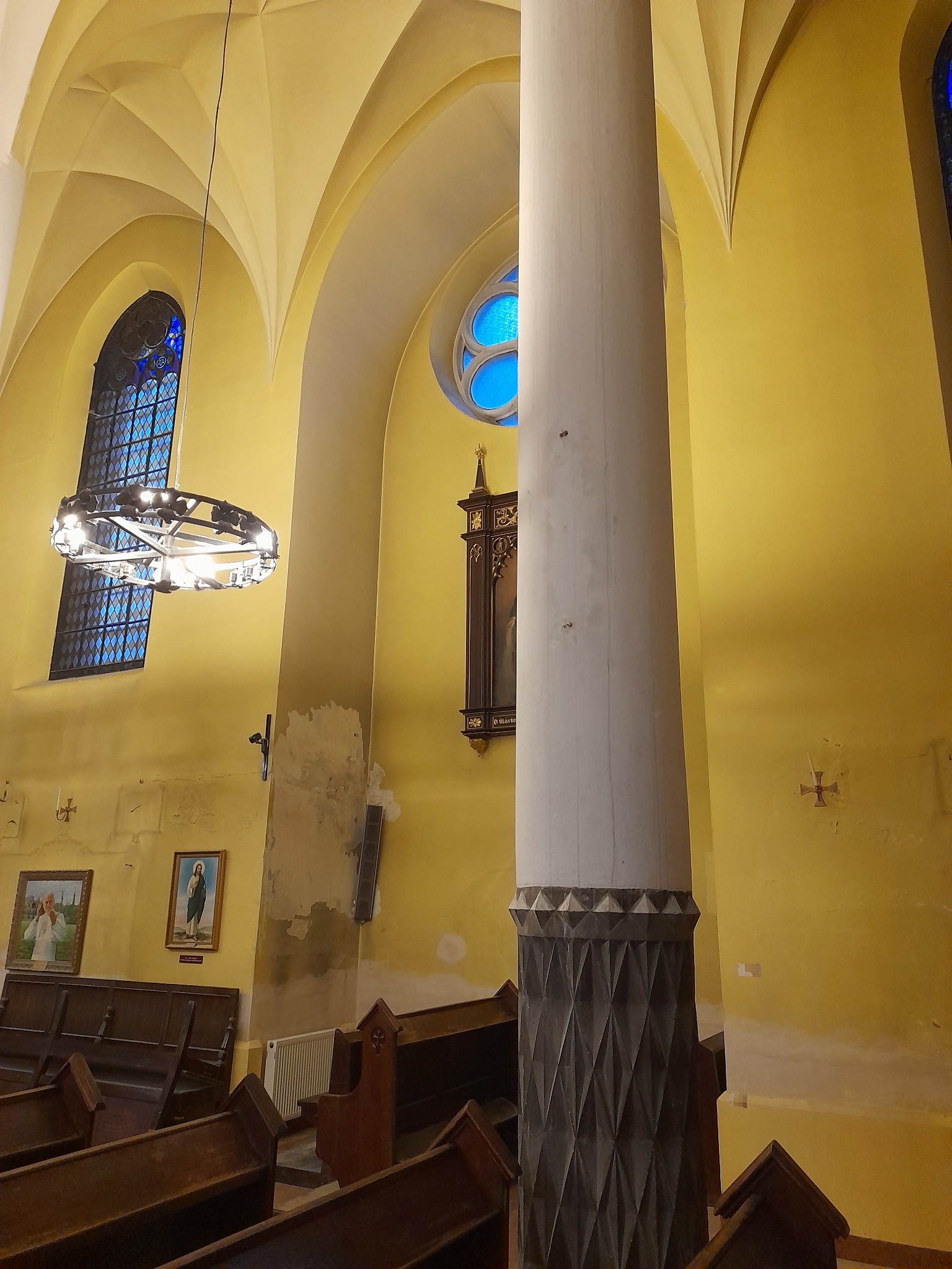 Wnętrze kościoła z żółtymi ścianami, drewnianymi ławkami i kolumną z dekoracyjnym wzorem u podstawy. Widoczne okna z witrażami i wiszący żyrandol.