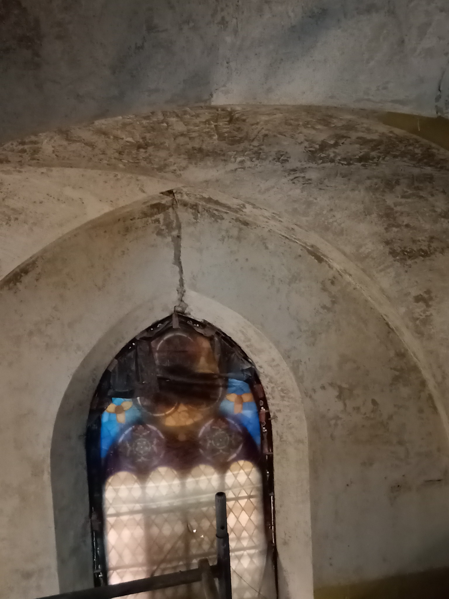 Krypta z łukowym sklepieniem i witrażem w oknie. Widoczna spękana, surowa powierzchnia ścian i sufitu. Witraż z motywami kwiatowymi w odcieniach niebieskiego i żółtego.