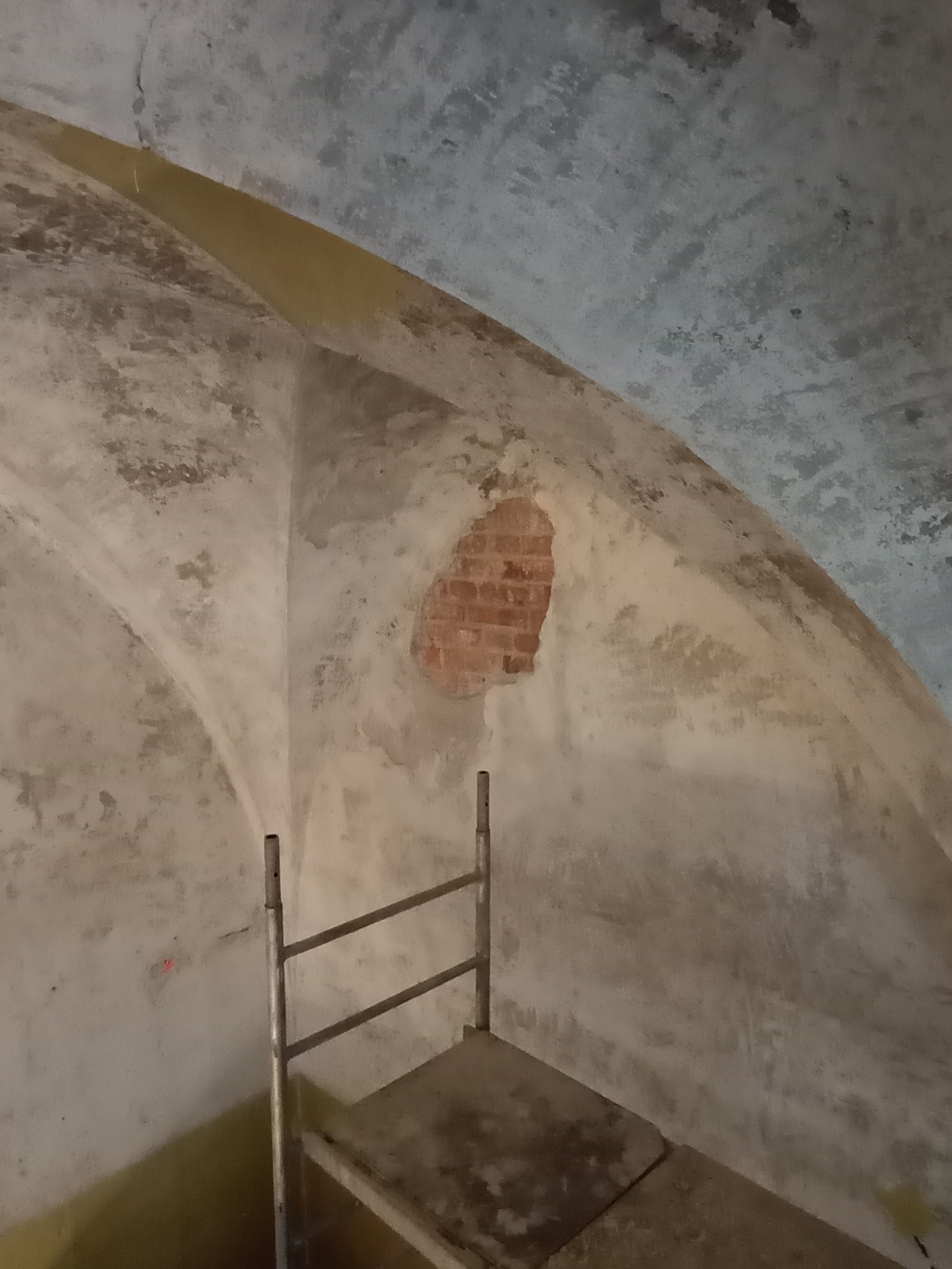 Klatka schodowa w trakcie remontu, widoczna ściana z fragmentem odsłoniętej cegły i metalowe rusztowanie. W tle sufit z widocznymi śladami wilgoci.