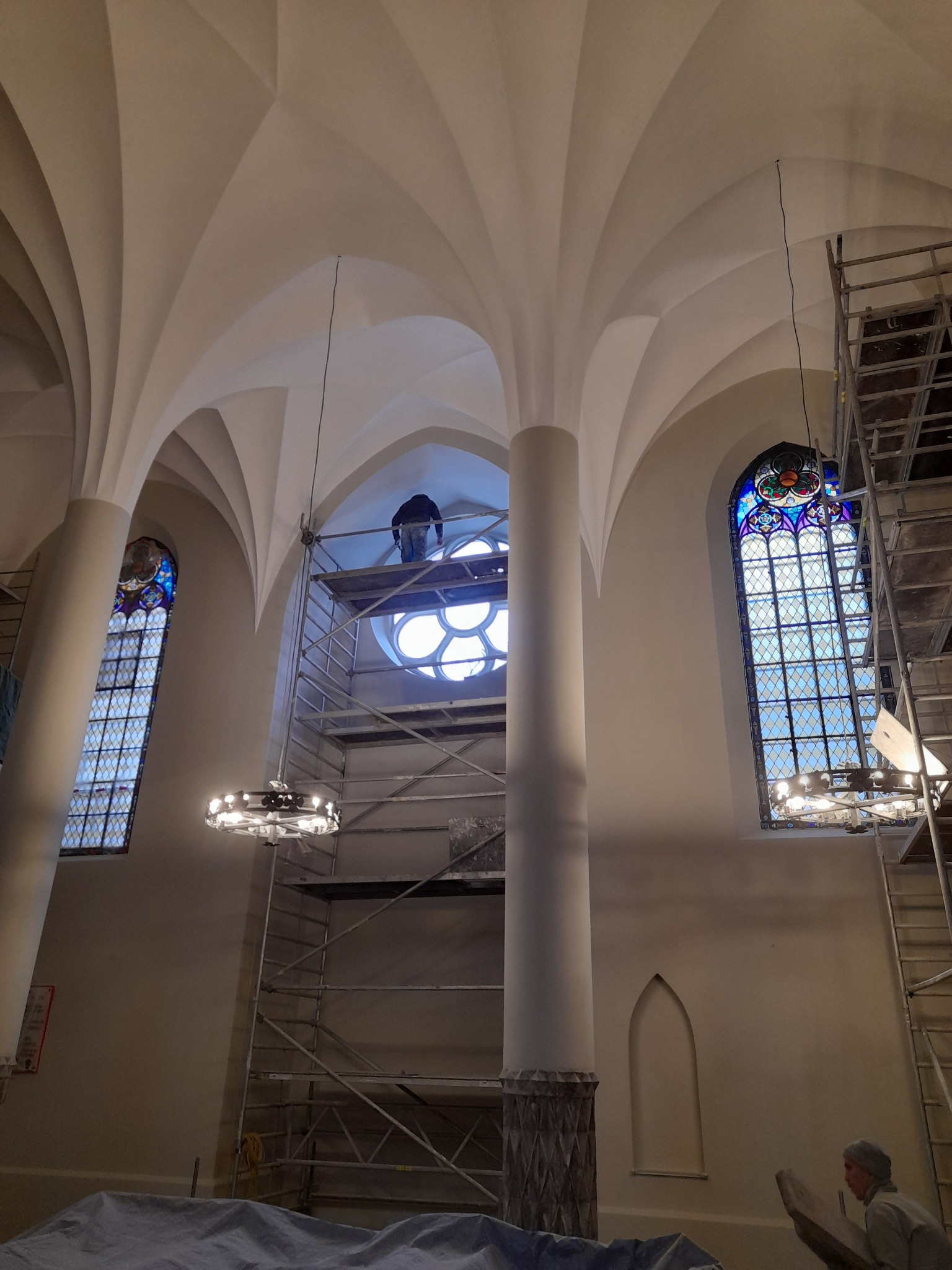 Wnętrze kościoła w trakcie renowacji: rusztowania, witraże, kolumny i sklepienie łukowe. Widoczny pracownik na rusztowaniu przy okrągłym oknie.