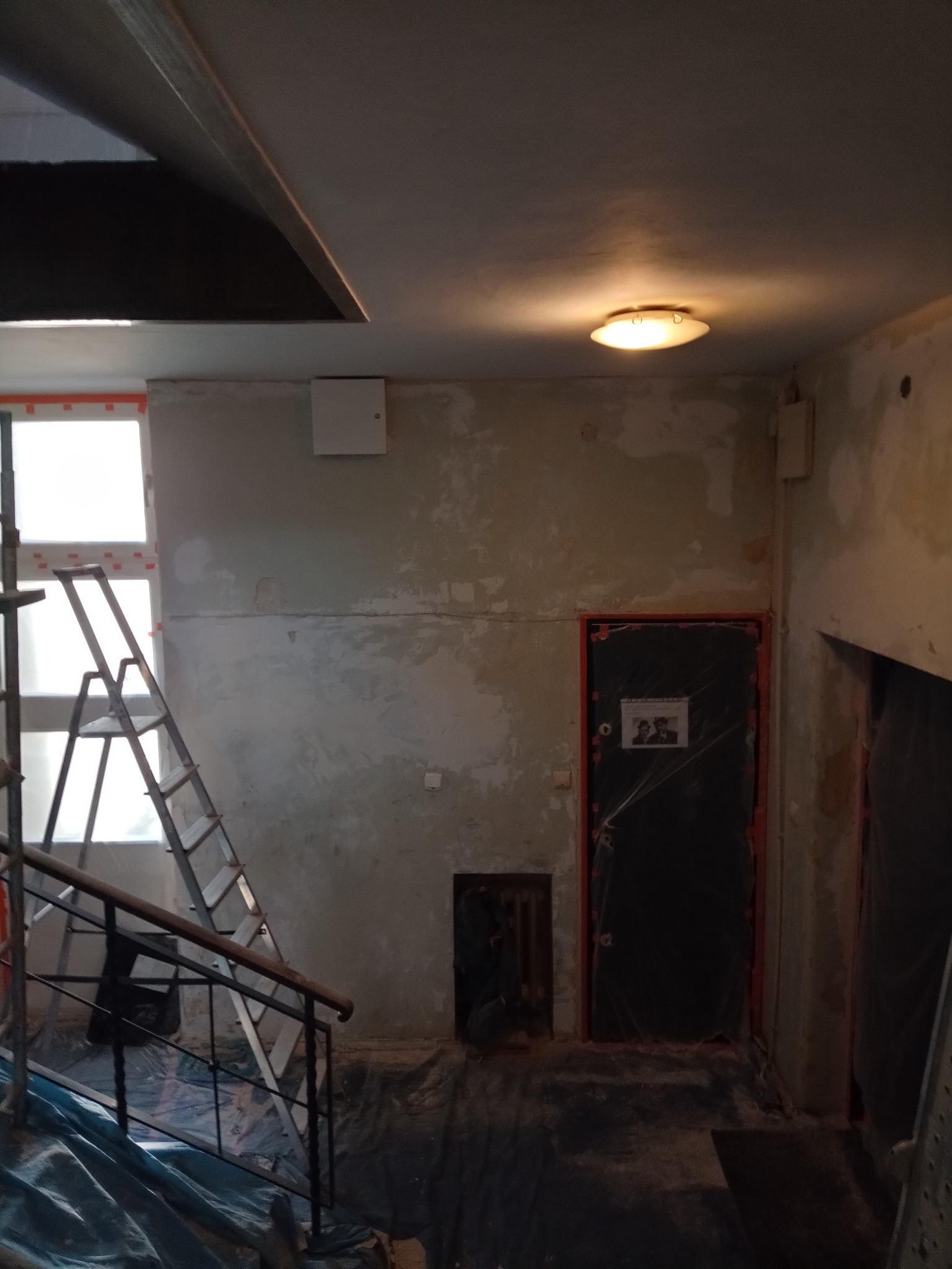 Klatka schodowa w trakcie remontu: ściany przygotowane do malowania, zabezpieczone folią drzwi, drabina oparta o okno, widoczne ślady po starych powłokach.