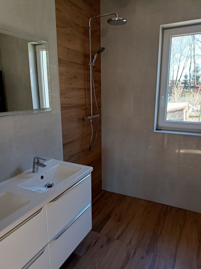 Nowoczesna łazienka z drewnopodobnymi płytkami, białą szafką pod umywalką i kabiną prysznicową. Widoczne okno z widokiem na zewnątrz i lustro.