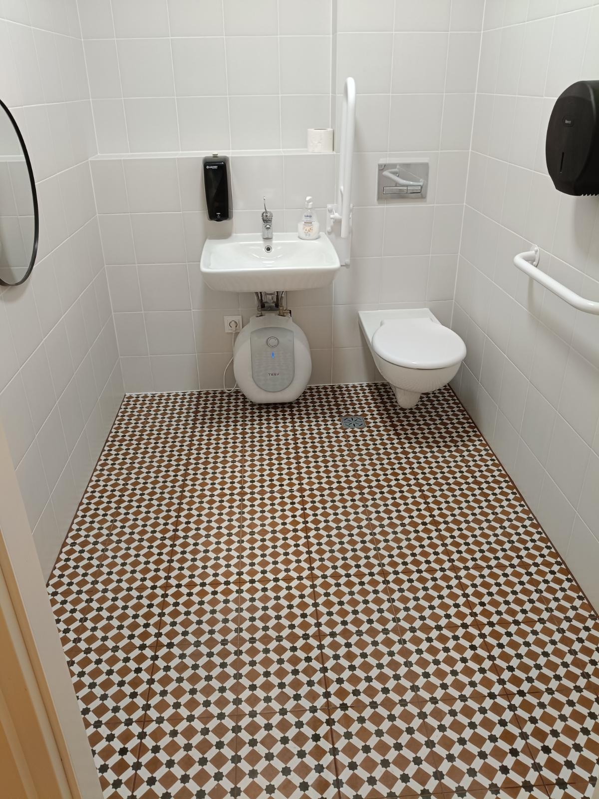 Wykończona łazienka z białą ceramiką, baterią, podgrzewaczem wody i wzorzystą, brązowo-białą podłogą. Udogodnienia dla osób z niepełnosprawnościami.
