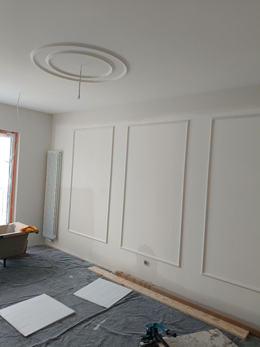 Białe ściany z dekoracyjnymi listwami i rozetą sufitową w trakcie prac wykończeniowych. Podłoga zabezpieczona folią, widoczne narzędzia i płytki.