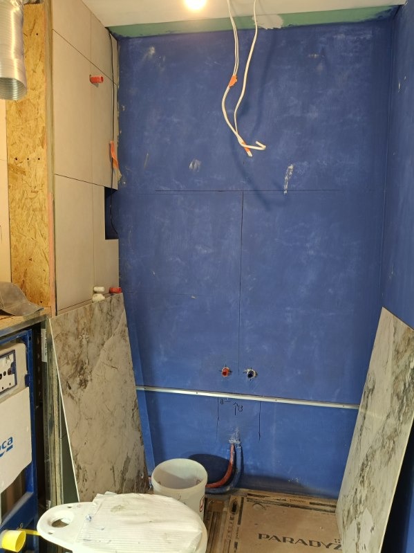 Niebieska ściana w trakcie przygotowania do wykończenia w łazience. Widoczne przewody, rury i płyty gipsowe. Częściowo ułożone płytki na ścianie.