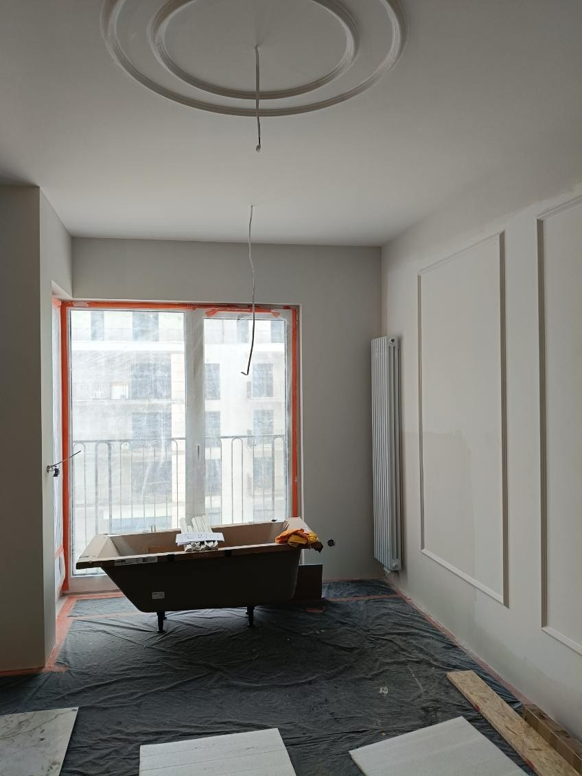 Łazienka w trakcie wykończenia: wanna w kartonie, zabezpieczona podłoga, listwy dekoracyjne na ścianie, sufit z rozetą. Widoczne przewody elektryczne.