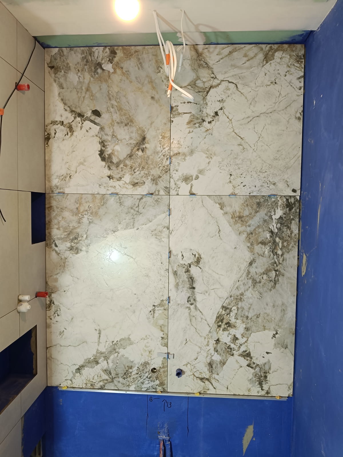 Układanie płytek imitujących marmur na ścianie w łazience. Widoczne niebieskie ściany i wystające kable. Etap prac wykończeniowych, z użyciem poziomowania płytek.