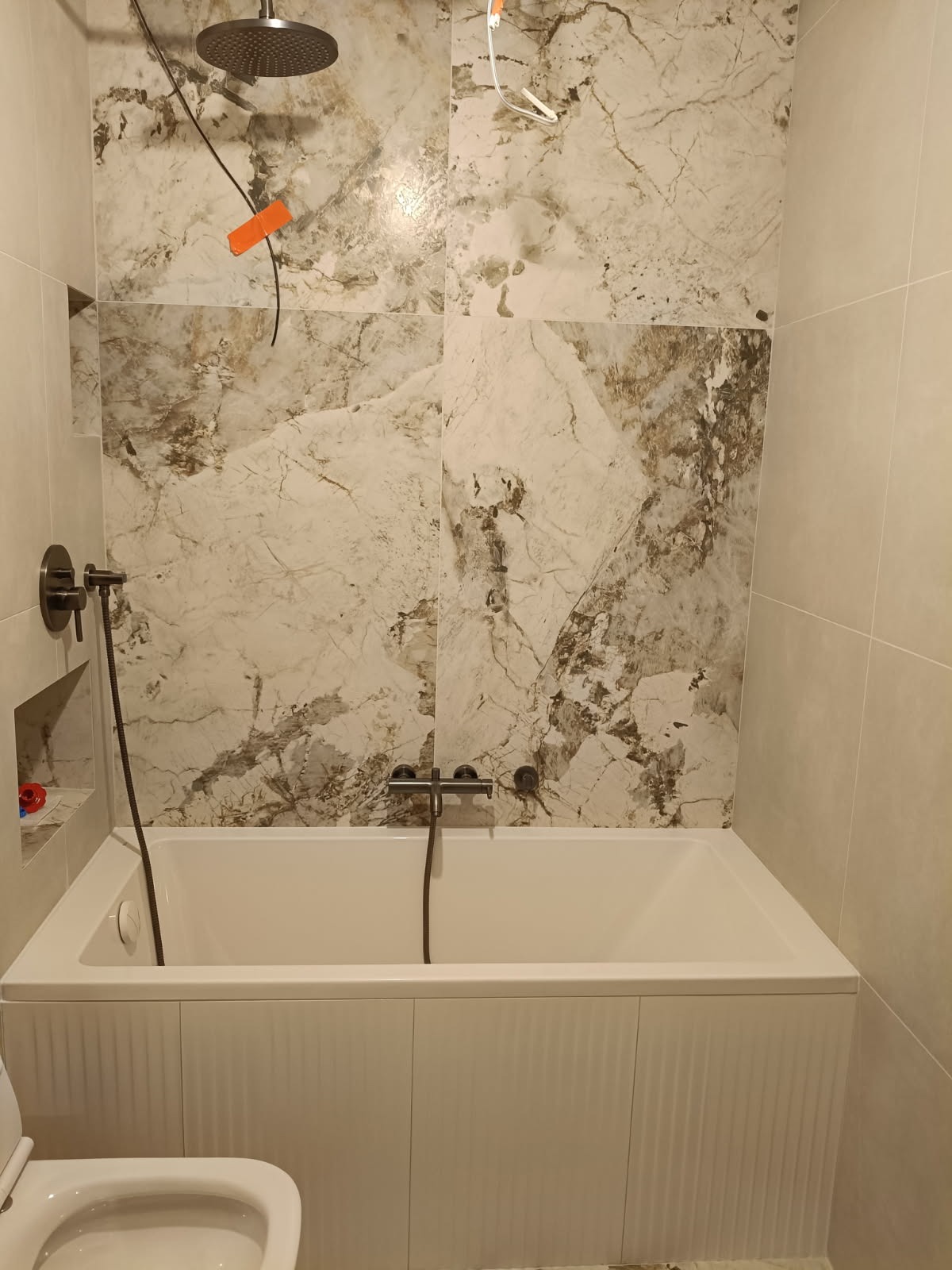 Wykończona łazienka z wanną obudowaną panelami, marmurowe płytki na ścianach, deszczownica i bateria w kolorze grafitowym. Widoczny fragment toalety.