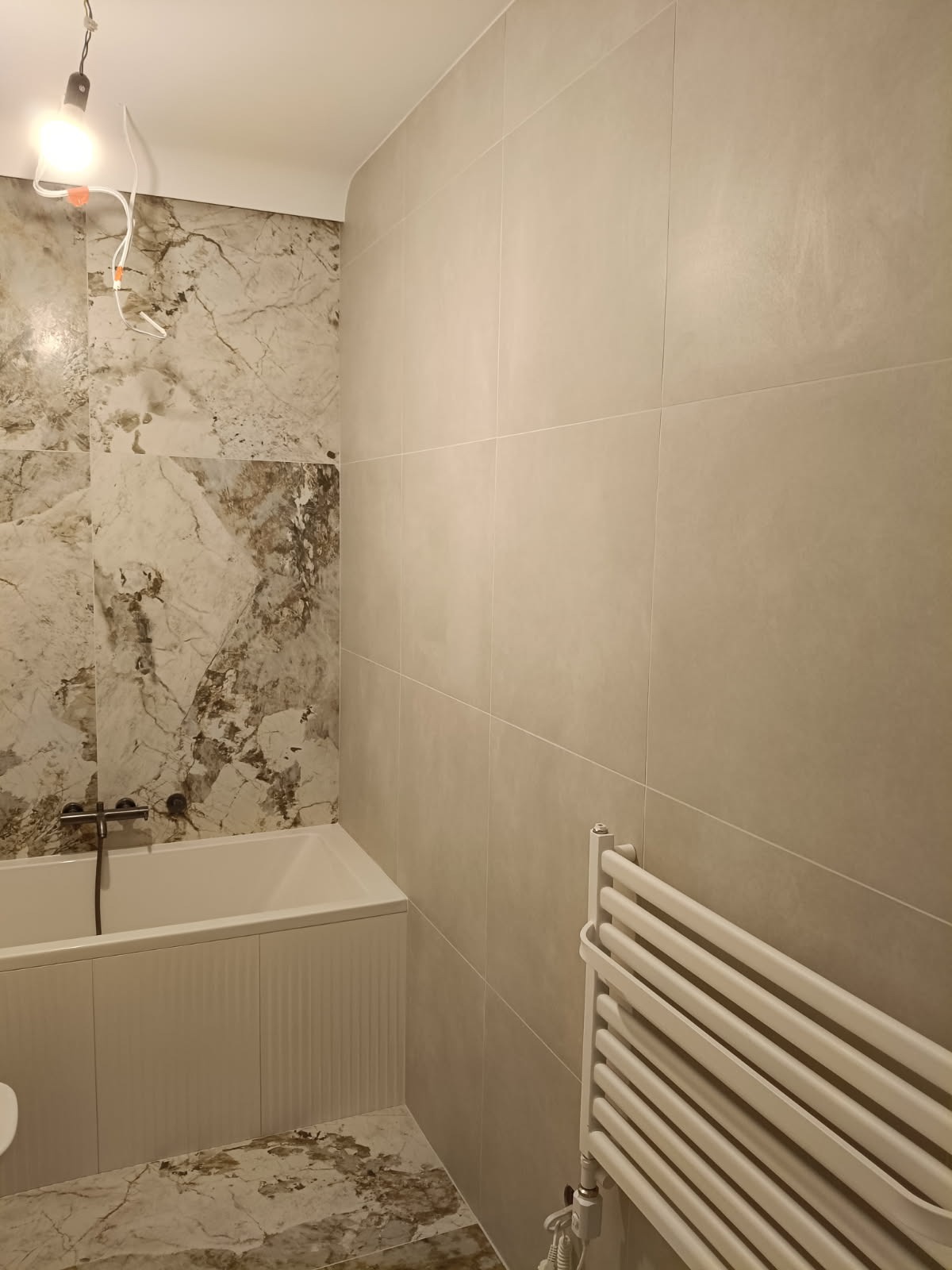 Wykończona łazienka z wanną, grzejnikiem drabinkowym i płytkami imitującymi marmur na ścianie. Widoczna żarówka na kablu. Minimalistyczny design.