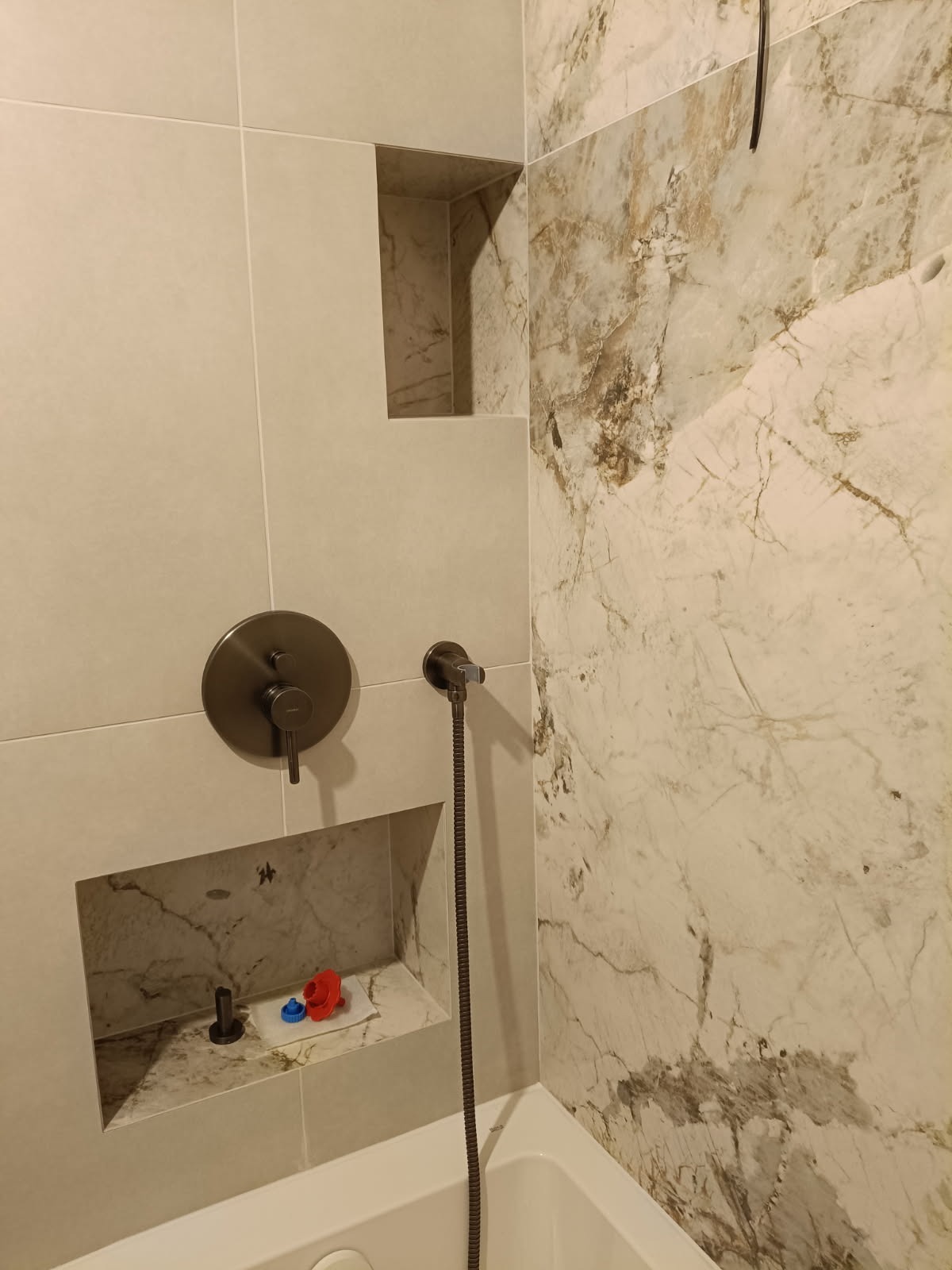 Wykończona łazienka z beżowymi i marmurowymi płytkami, wnękami w ścianie, baterią prysznicową w kolorze ciemnego brązu i białą wanną. Widok z bliska na detale.