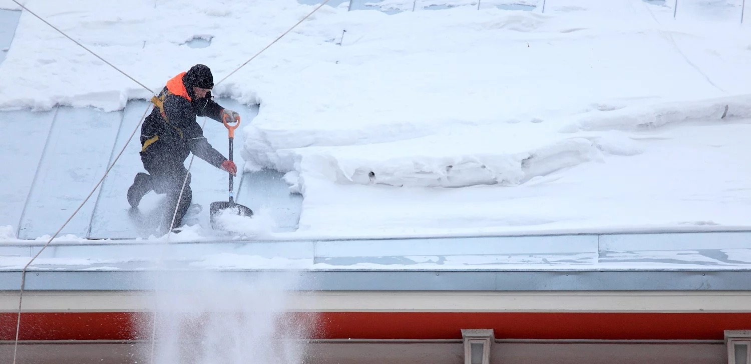 Pracownik w kombinezonie odśnieża płaski dach budynku za pomocą łopaty, odrzucając śnieg na bok. Widoczne liny zabezpieczające i fragment elewacji.